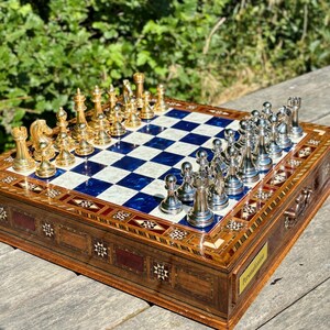 A chess set with a blue and white checkered board and gold and silver pieces is displayed on a wooden surface, with a natural outdoor background.