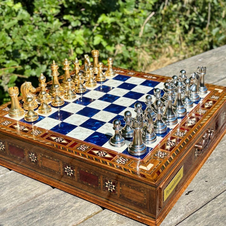 A chess set with a blue and white checkered board and gold and silver pieces is displayed on a wooden surface, with a natural outdoor background.
