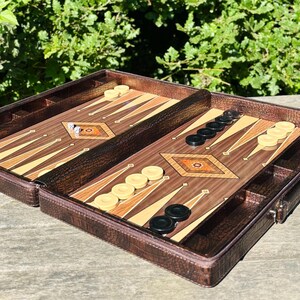 An open brown leather game case containing a backgammon game board and pieces, placed on a wooden surface with greenery in the background.