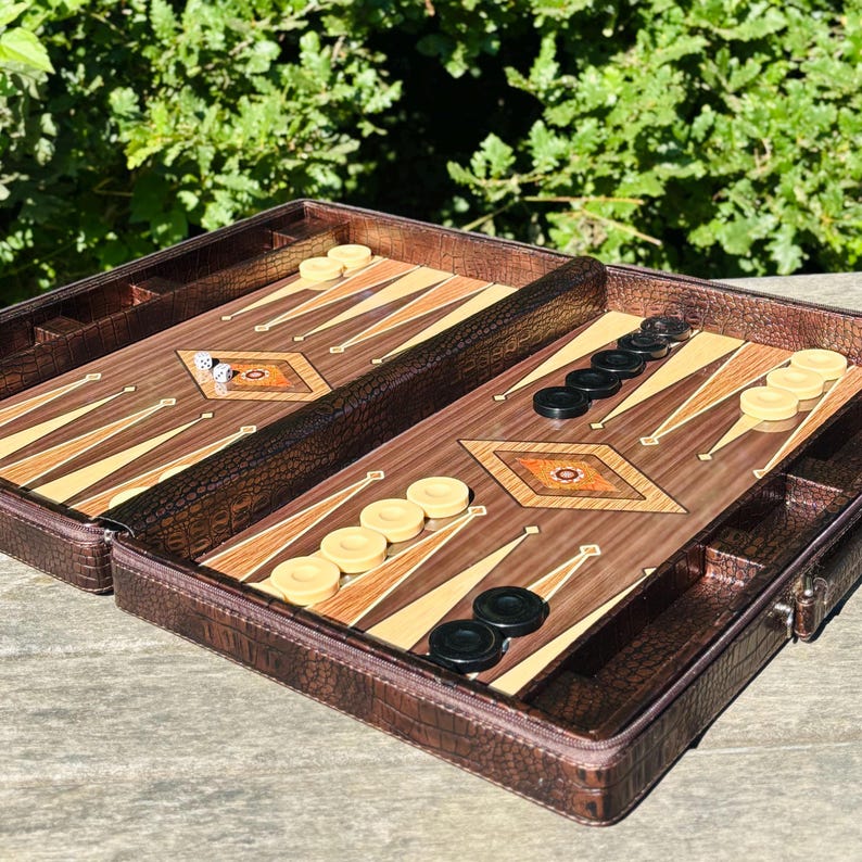 An open brown leather game case containing a backgammon game board and pieces, placed on a wooden surface with greenery in the background.