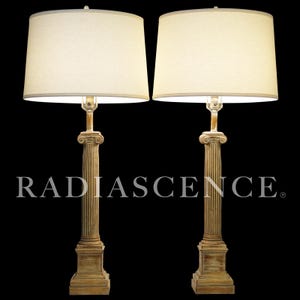 May include: A pair of table lamps featuring beige shades and column-style bases. The bases have a distressed, aged beige finish with vertical grooves and decorative details. The lamps are set against a black background, with the word "RADIASCENCE" printed on the base.