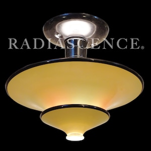 May include: A ceiling light fixture with a polished chrome canopy and a two-tiered, pale yellow glass shade featuring dark brown trim. The word RADIASCENCE is printed in white capital letters across the upper portion of the light fixture.