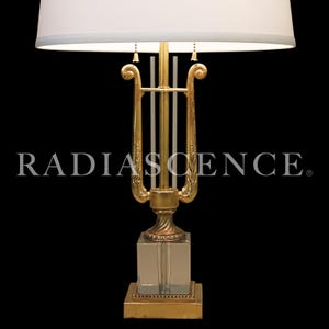Op de afbeelding: Een goudkleurige tafellamp met een witte lampenkap. De lamp heeft een lier-vormig ontwerp en een heldere, vierkante basis. De tekst "RADIASCENCE" is zichtbaar op de lamp.