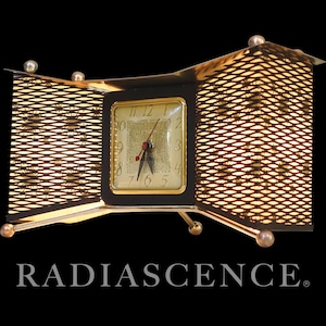 Puede incluir: Un reloj vintage con esfera dorada y marco negro, ubicado dentro de una estructura metálica rectangular de tejido abierto. El reloj tiene manecillas doradas y un segundero rojo. La palabra "RADIASCENCE" se muestra debajo.