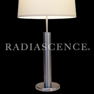 May include: A table lamp with a brushed metal cylindrical base and a circular stand, topped with a wide, off-white drum shade. The word RADIASCENCE is printed in white capital letters across the center of the lamp against a black background.