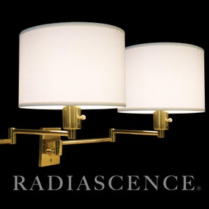 May include: Two wall-mounted lamps with white cylindrical shades and brass-colored articulating arms. The lampshades have a light gray trim. The base of the lamp is rectangular. The word "RADIASCENCE" is printed at the bottom.