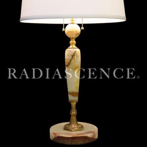 May include: A table lamp with a white drum shade and a tall, slender body made of polished cream and brown onyx stone. The lamp features gold-toned metal accents, a round onyx base, and two pull chains. The word RADIASCENCE is printed in white.
