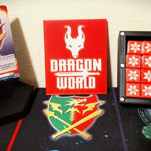 May include: A display of gaming accessories featuring a card, a red sign with "DRAGON WORLD" text, and a container of red dice. The card has a dragon and shield graphic. The dice are red with white snowflake designs.