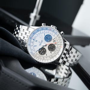 Peut inclure: Une montre Breitling Navitimer argentée avec un cadran blanc, plusieurs sous-cadrans et un affichage de la date. La montre a un bracelet en acier inoxydable et est présentée sur une surface réfléchissante.