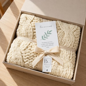 May include: A cream-colored chunky knit cable blanket folded inside a beige gift box. A white card with a green leaf illustration and the text "There are no words. Sending you love, comfort and strength during this difficult time" is tied to the blanket with a beige ribbon. A small white tag with the text "B LUXE Luxury & style delivered" is attached.