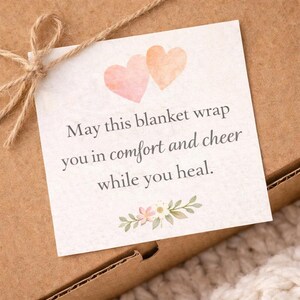 Puede incluir: Una tarjeta cuadrada con el texto "May this blanket wrap you in comfort and cheer while you heal" est&aacute; atada a una caja de cart&oacute;n marr&oacute;n con hilo. La tarjeta tiene dos corazones de acuarela y detalles florales.