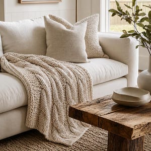 May include: A cream-coloured cable knit throw blanket draped over a light beige sofa. The sofa features two square throw pillows in matching cream tones. A textured beige area rug covers the floor, and a wooden coffee table sits in the foreground.