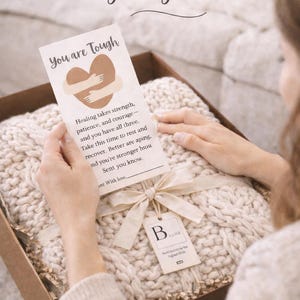 Puede incluir: Una caja de regalo que contiene una manta de punto grueso de color crema y una tarjeta que dice "Thinking of You During Tough Times". La tarjeta tiene un gr&aacute;fico de coraz&oacute;n y texto de apoyo. Cinta y etiqueta color crema.