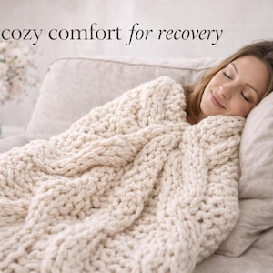 Puede incluir: Una persona descansa en un sof&aacute;, cubierta por una gruesa manta de punto color crema. La manta tiene un dise&ntilde;o de punto trenzado grueso. El texto "cozy comfort for recovery" est&aacute; en la parte superior de la imagen. Un jarr&oacute;n con flores est&aacute; en una mesa auxiliar.