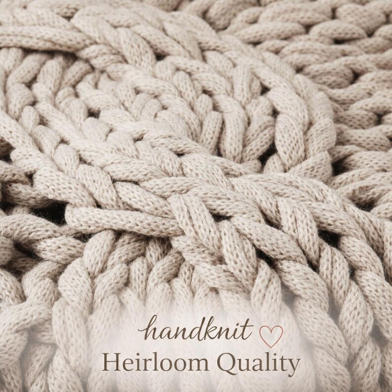 Puede incluir: Primer plano de una manta beige tejida a mano con un grueso patr&oacute;n de punto trenzado. Las palabras "handknit" y "Heirloom Quality" est&aacute;n escritas en cursiva en la parte inferior de la imagen.