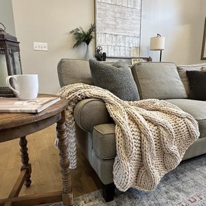 Puede incluir: Una acogedora sala de estar con un sofá gris claro adornado con cojines y una manta beige de punto grueso. Una mesa auxiliar de madera sostiene una taza blanca y una foto enmarcada. Una linterna vintage y un espejo decorativo completan la escena.