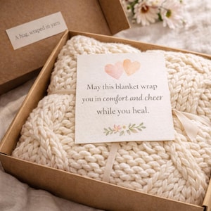 Puede incluir: Una manta de punto grueso de color crema se presenta en una caja marr&oacute;n. Una peque&ntilde;a tarjeta con el texto "May this blanket wrap you in comfort and cheer while you heal" est&aacute; atada a la manta. La caja tambi&eacute;n tiene una etiqueta que dice "A hug, wrapped in yarn."