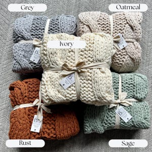 May include: A collection of chunky knit blankets in various colors, including grey, ivory, rust, oatmeal, and sage. Each blanket is neatly folded and tied with a ribbon and a tag. The blankets are displayed on a neutral-toned surface. Realtor Closing gift ideas for cozy home decor keepsake