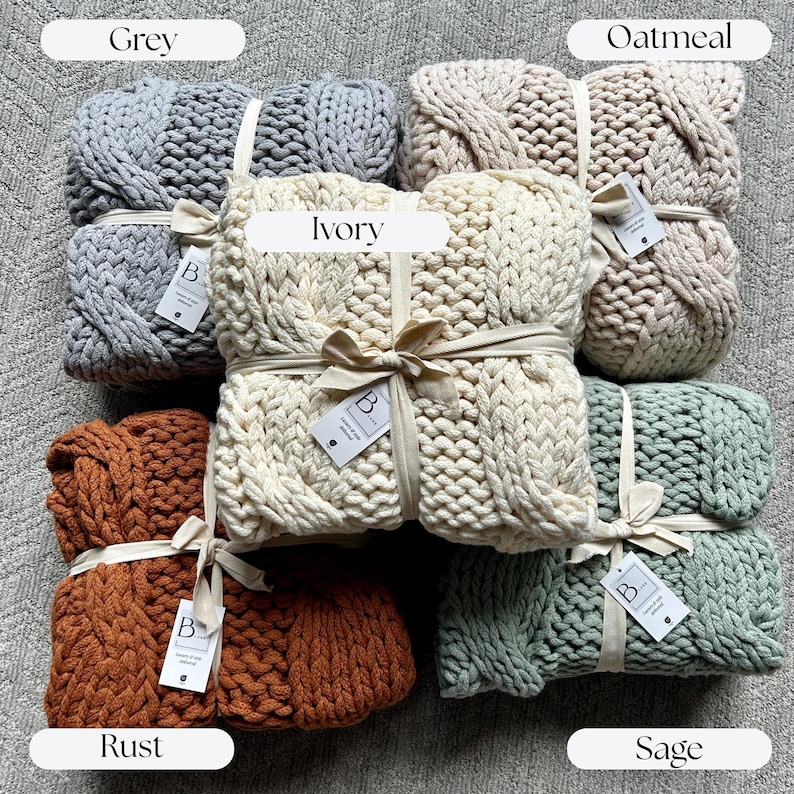 May include: A collection of chunky knit blankets in various colors, including grey, ivory, rust, oatmeal, and sage. Each blanket is neatly folded and tied with a ribbon and a tag. The blankets are displayed on a neutral-toned surface. Realtor Closing gift ideas for cozy home decor keepsake