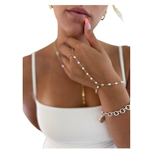 May include: Gold-toned hand chain bracelet with small white pearls and a ring. A silver bracelet with a heart charm is also visible. The jewelry is worn on a person's hand and wrist.