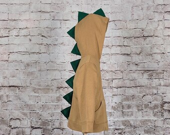 May include: A tan dinosaur-themed hoodie with a hood and a zipper. The hoodie features dark green triangular spikes along the back and hood, resembling a dinosaur's spine. The hoodie is displayed on a stand against a brick wall.