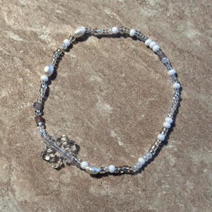 Könnte beinhalten: Ein filigranes Armband mit weißen und transparenten Perlen, durchsetzt mit kleinen silbernen Perlen und einigen größeren, unregelmäßigen weißen Perlen. Ein klarer, schneeförmiger Anhänger sorgt für einen Hauch von Verspieltheit. Das Armband hat einen Umfang von etwa 18 cm.