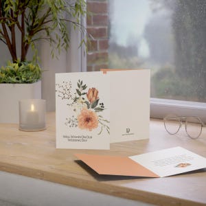 Op de afbeelding: Trouw wenskaart met bloemmotief in perzik, wit en groen. De kaart leest "Well Wishes On Our Wedding Day". De kaart is open en onthult een bijpassend perzik kleurig interieur.