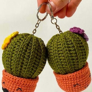 Puede incluir: Dos llaveros de cactus hechos a mano con ganchillo. Los cactus son verdes con detalles de flores amarillas y moradas, y están colocados en macetas naranjas con caras sonrientes. Cada llavero está sujeto a un llavero plateado.