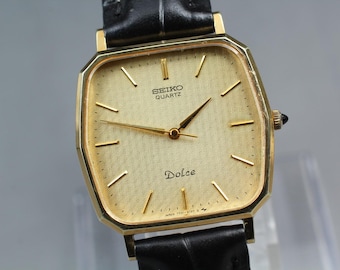 May include: A gold-toned Seiko Quartz Dolce watch with a rectangular face and black leather band. The watch face has gold hands and markers, with the word "Dolce" printed at the bottom. The watch is a classic timepiece.