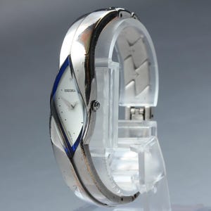 May include: A silver-toned Seiko wristwatch with a white face and blue accents. The watch has a unique, curved design and is displayed on a clear stand. The watch face has small, silver-toned markers.