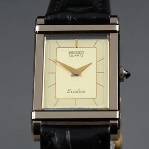 May include: A rectangular Seiko Quartz Excaline wristwatch featuring a champagne-colored dial with gold-tone hands and hour markers. The watch has a metallic frame and is attached to a black textured leather strap with a visible winding crown.