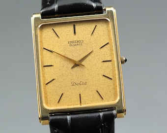 May include: A gold-toned Seiko Quartz Dolce watch with a rectangular face and black leather band. The watch face is gold with black markers and hands. The word "Dolce" is printed on the watch face.
