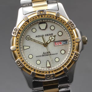 Puede incluir: Un reloj Alba Aqua Gear plateado y dorado con esfera blanca. El reloj muestra la fecha "DOM 3" y es resistente al agua hasta 20 bar. Tiene un bisel dorado con marcas negras y una correa de metal plateada y dorada.