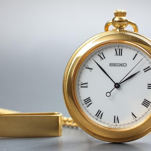 May include: A gold-coloured Seiko pocket watch with a white face and black Roman numerals. The watch has a matching gold chain and a rectangular fob. The watch face displays the Seiko logo.