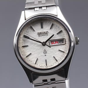 May include: A silver Seiko Quartz watch with a textured white face. The watch displays the day and date, with "SUN 5" visible. The watch has black hands and markers, and a silver band. The watch is a vintage style.