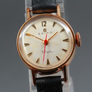 May include: A vintage Seiko wristwatch with a gold-tone case and a cream-colored face. The watch features gold hands, hour markers, and the text "Seiko" and "Pimple". The watch has a black leather strap.