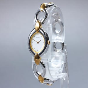 May include: A Seiko women's wristwatch with a silver and gold-tone oval watch face and a white dial. The watch has a linked bracelet with gold-tone accents. The watch is displayed on a clear stand.