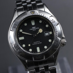 Puede incluir: Un reloj Technos Quartz Super Diver de acero inoxidable con esfera negra. El reloj presenta agujas e indicadores luminosos, una ventana de fecha y un bisel giratorio. La correa del reloj es de acero inoxidable.