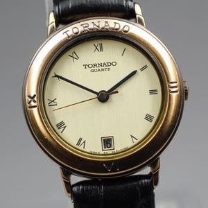 Könnte beinhalten: Eine goldfarbene Tornado Quartz Armbanduhr mit cremefarbenem Zifferblatt und schwarzen Zeigern. Die Uhr verfügt über römische Ziffern, eine Datumsanzeige und ein schwarzes Lederarmband. Auf dem Zifferblatt steht das Wort "Tornado" über "Quartz".