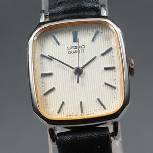 Könnte beinhalten: Eine Vintage Seiko Quartz-Uhr mit silberfarbenem Gehäuse und schwarzem Lederarmband. Das Zifferblatt ist weiß mit schwarzen Zeigern und Markierungen, und der Text "SEIKO QUARTZ" ist sichtbar. Die Uhr hat eine quadratische Form mit abgerundeten Ecken.