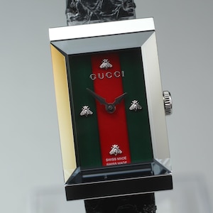 Könnte beinhalten: Rechteckige Gucci-Uhr mit grün-rotem Streifenzifferblatt, silbernen Akzenten und einem schwarzen Lederarmband. Das Zifferblatt zeigt das Gucci-Logo und Bienenmotive. Swiss Made.