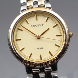 Könnte beinhalten: Eine Lucent Seiko Armbanduhr mit goldfarbener Lünette und silberfarbenem Armband. Das Zifferblatt ist cremefarben mit goldfarbenen Stundenmarkierungen und Zeigern. Das Wort "LUCENT" ist oben und unten auf der Lünette zu sehen.