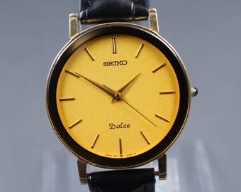 May include: A gold-toned Seiko Dolce watch with a black leather band. The watch face is a vibrant gold color with gold hands and markers. The watch has the brand name "SEIKO" and the word "Dolce" printed on the face.