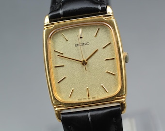May include: A gold-toned Seiko wristwatch with a rectangular face and a black leather band. The watch face is a light gold color with gold hands and markers. The word "SEIKO" is printed on the watch face.