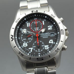 May include: A silver Seiko Chronograph watch with a black dial, featuring white numerals and red accents. The watch has multiple sub-dials, a date display, and a stainless steel band. The watch is water resistant to 100 meters.