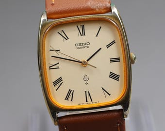 May include: A vintage Seiko Quartz watch with a gold-toned rectangular case and a cream-colored dial. The dial features black Roman numeral hour markers and black hands. The watch has a brown leather strap.