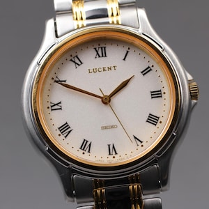Puede incluir: Reloj de pulsera Lucent Seiko plateado y dorado. El reloj tiene una esfera blanca con números romanos negros y agujas doradas. La correa es plateada con detalles dorados. La esfera mide aproximadamente 3,2 cm de diámetro.
