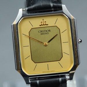 May include: A Credor Seiko wristwatch with a square silver-tone case and a two-tone yellow and gold dial. The watch features thin black hour markers, gold hands, and a black leather strap. The text Credor and Seiko are printed on the dial.