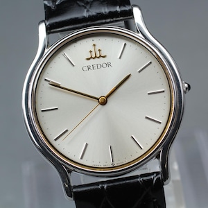 May include: A Credor wristwatch featuring a round silver-toned dial with gold-colored hands and hour markers. The watch has a polished silver-tone metal case and a black textured leather strap. The Credor logo is printed in gold at the top center.