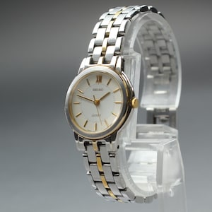 Puede incluir: Un reloj Seiko Excelsior plateado y dorado con esfera blanca y agujas doradas. El reloj tiene una correa de eslabones plateada y dorada. El reloj se exhibe sobre un soporte de plástico transparente sobre un fondo gris.